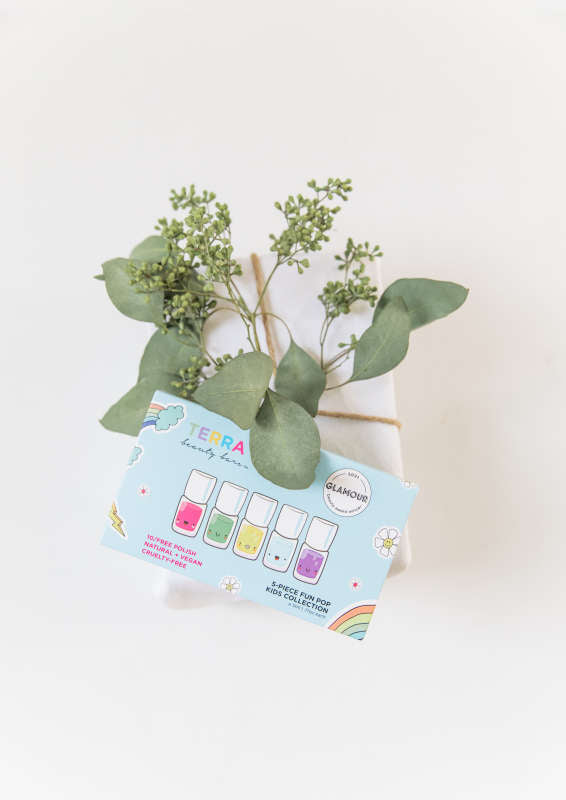 A potted plant with green leaves sits on a white surface. In front of it, a colorful card features Terra Beauty Bars Kids 5-Piece Fun Pop Nail Collection, offering vibrant, vegan, cruelty-free polish shades. The softly lit background remains plain and serene.