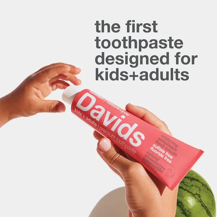 A child’s hand and an adult’s hand hold a red tube of Davids Kids + Adults Premium Toothpaste; text above reads, “the first fluoride free toothpaste designed for kids and adults.”.