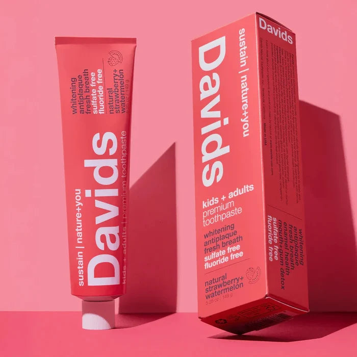 A pink tube and matching box of Davids Kids + Adults Premium Toothpaste with white lettering, shown against a pink backdrop. The packaging emphasizes that it is a fluoride-free toothpaste crafted with natural ingredients and offers a tasty strawberry flavor suitable for both children and adults.