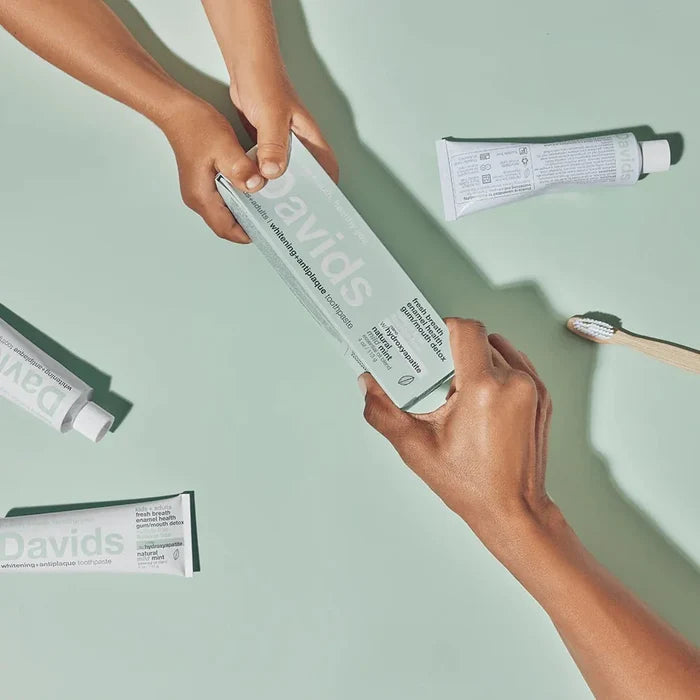 Two hands exchange a tube of Davids Hydroxi Whitening + Enamel Health Toothpaste with nano hydroxyapatite for Kids and Adults on a light green surface, surrounded by more fluoride-free toothpaste tubes and a toothbrush, emphasizing the brand’s dedication to natural whitening.