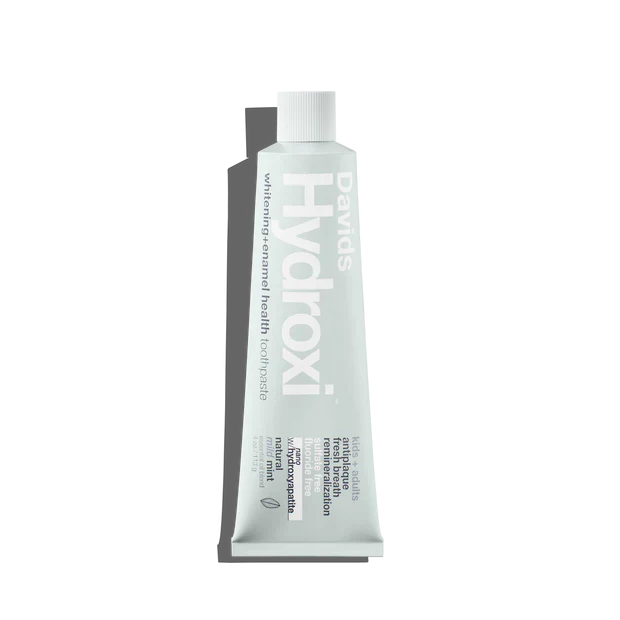 A white tube of Davids Hydroxi Whitening + Enamel Health Toothpaste w/nano hydroxyapatite, Kids+Adults stands upright on a light background, with the text and branding displayed vertically on the tube. This fluoride-free toothpaste uses nano hydroxyapatite to naturally whiten teeth and support enhanced enamel health.