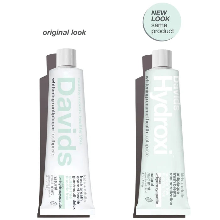 Two toothpaste tubes stand upright side by side. The left tube is labeled “Davids,” and the right tube, labeled “Hydroxi Whitening + Enamel Health Toothpaste w/nano hydroxyapatite, Kids+Adults,” features a “NEW LOOK same product” sticker above it, highlighting the packaging change for this fluoride-free Davids toothpaste formulated with nano hydroxyapatite for natural whitening.