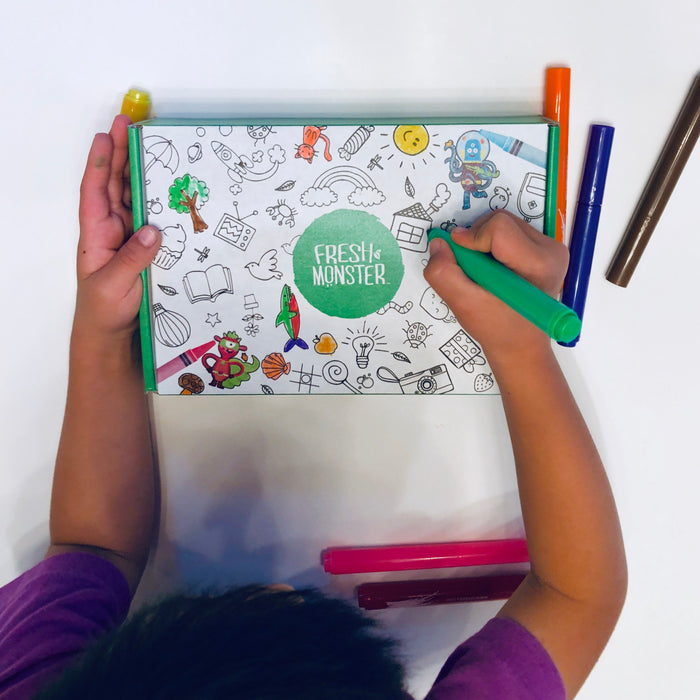 A child joyfully colors a Fresh Monster activity booklet using a green marker from their coloring box, surrounded by black-and-white cartoon and doodle illustrations. The table is scattered with red, orange, and brown markers while enjoying a Kids & Baby Gift Set by Fresh Monster.