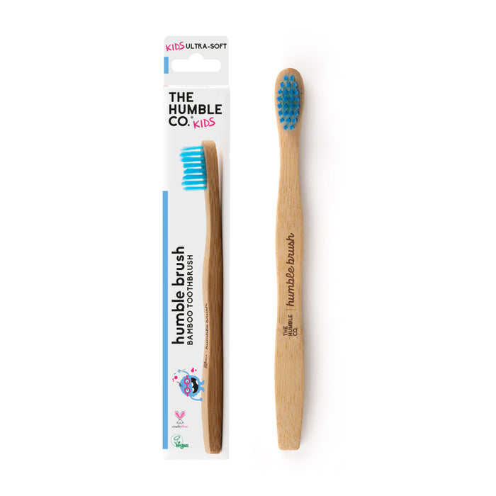 A Kids Bamboo Toothbrush - Ultra Soft from The Humble Co. rests beside its packaging. It features blue nylon bristles and a wooden handle with humble brush printed, making it an eco-friendly choice for young ones.