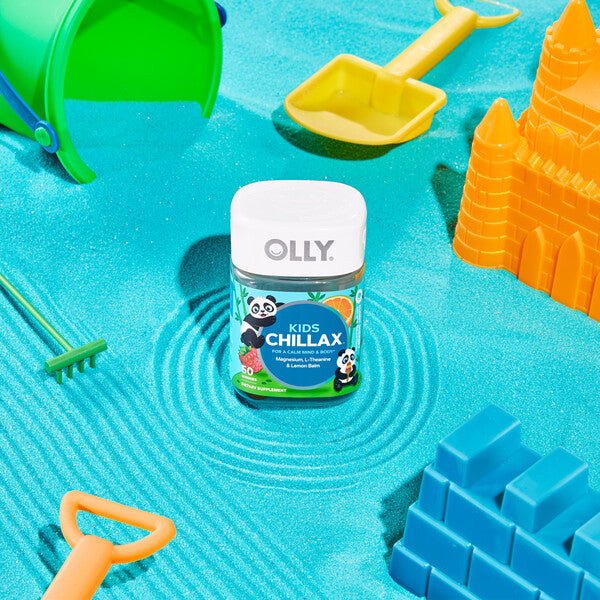 A container of OLLYs Kids Chillax calming gummies sits on bright blue sand with a green bucket, yellow shovel, and orange sandcastle mold, creating a lively summery scene. These gluten-free treats promise peace and playfulness all day.