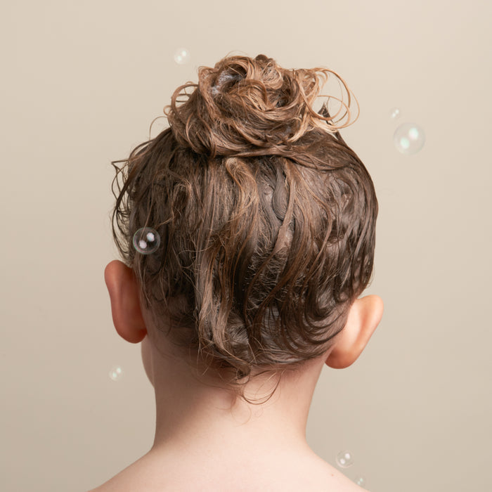 Rear view of a person with wet hair styled in a messy bun against a beige background. Several soap bubbles float around their head, suggesting they used the For Tangles Conditioner Bar by NOT!CE Hair Co for an effortlessly playful look and essential kids hair care routines.