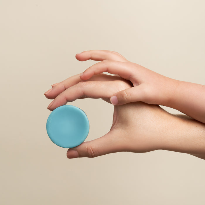 Two hands gently hold a circular light blue For Tangles Conditioner Bar by NOT!CE Hair Co against a neutral background, highlighting the symmetry and balance—ideal for kids hair care routines.