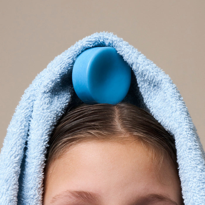 The NOTICE Hair Co Kids Conditioner Bar - For Tangles, in blue, sits atop a child's wet brown hair, partially wrapped in a light blue towel against a plain beige background.
