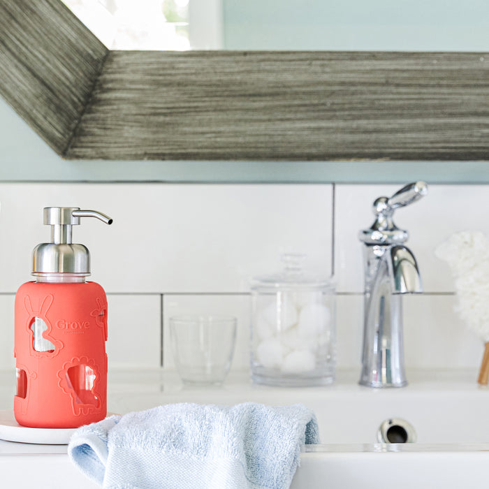 A bathroom sink features a silver faucet and mirrored backsplash. On the counter, theres a Grove Co. Kids Foaming Hand Soap Glass Dispenser with red and white animal designs, perfect for kids, accompanied by a light blue towel, a glass jar of cotton balls, and a tumbler in an eco-friendly setup.