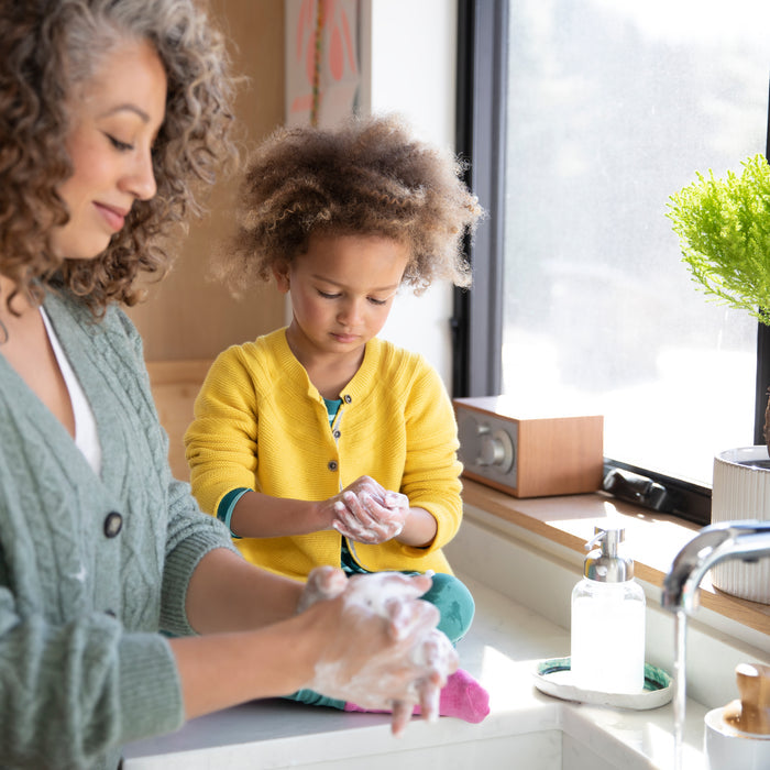 A woman and child wash their hands using Grove Co. Kids Foaming Hand Soap in a reusable glass dispenser. The child, wearing a yellow sweater, is focused and enjoys the moment while the woman smiles. Nearby, a plant and soap dispenser adorn the windowsill.
