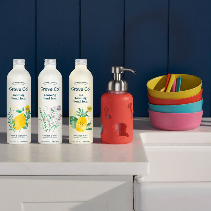 Three Grove Co. Kids Foaming Hand Soap bottles in Lavender & Eucalyptus, Pineapple, and Fresh Citrus sit with a red pump on a bathroom counter. The glass dispenser aims to reduce plastic waste, while colorful bowls are stacked against the blue wall backdrop.
