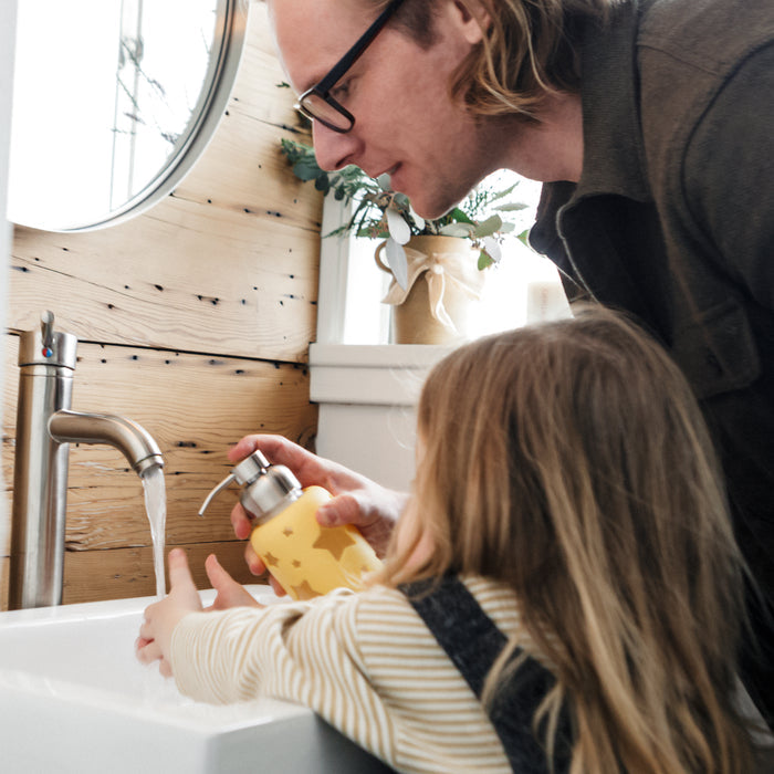A man and a child wash hands at a sink with Grove Co. Kids Foaming Hand Soap Refill. The man holds a yellow soap dispenser while the child reaches for water in their bathroom with a wooden wall and potted plant, enjoying soap made from plant-based ingredients.