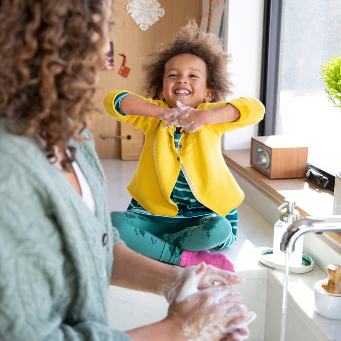 A young child in a yellow cardigan and green pants joyfully raises their arms by a window as a curly-haired woman uses Grove Co.s Kids Foaming Hand Soap Refill. Natural light illuminates the scene, highlighting the plastic-free packaging.