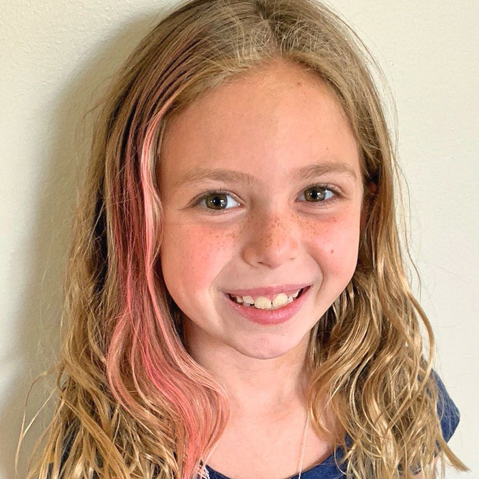 A young girl with long, wavy blonde hair adorned with a pink streak from Fresh Monsters Kids Hair Chalk smiles at the camera. She wears a dark blue top and stands against a light-colored background.