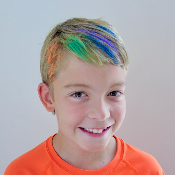 A child with short blonde hair beams with joy, their locks transformed by Fresh Monsters hypoallergenic Kids Hair Chalk-Temporary in vibrant blue, green, and orange. They wear an orange shirt against a white background, highlighting the fun of temporary hair color.