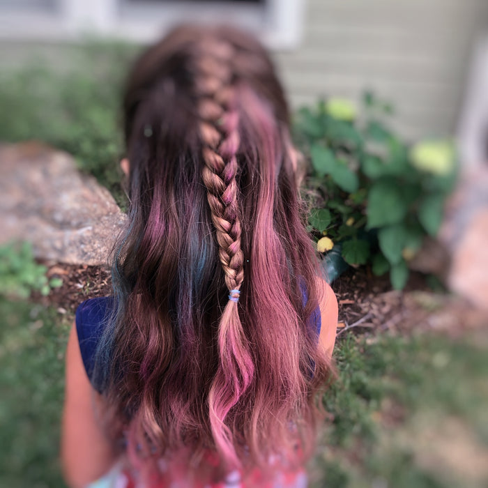 A young girl with long, wavy hair is styled in a vibrant braid using Fresh Monsters Kids Hair Chalk-Temporary in shades of pink, blue, and brown. Standing outdoors among greenery and rocks, her temporary hair color beautifully complements the natural setting.