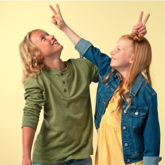 Two smiling children pose together. The one in a green shirt laughs while the other, in a yellow dress and denim jacket, holds up bunny ears—radiating the same happy energy as taking MegaFood Kids Multi Gummies daily!.