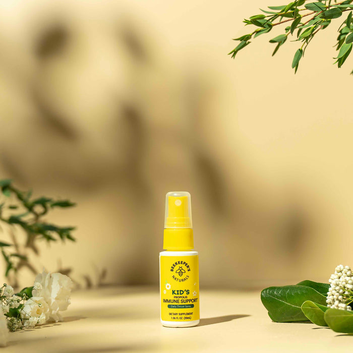 A yellow Kids Propolis Immune Support Throat Spray bottle from Beekeepers Naturals sits on a beige background with green leaves and white flowers, evoking a natural setting. The bee logo indicates propolis as the key ingredient for immune support.