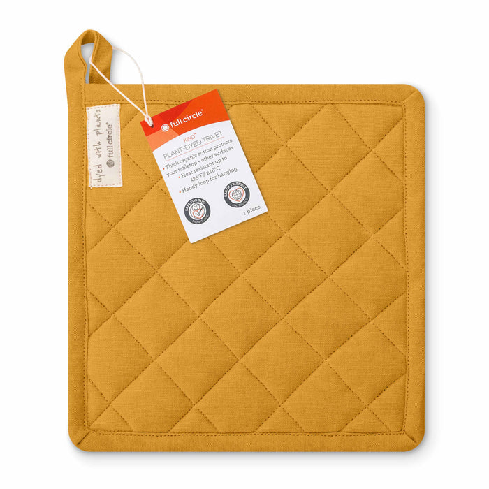 The Kind Plant Dyed Trivet by Full Circle is a square mustard-yellow quilted pot holder made from organic cotton with a stitched diamond pattern and a hanging loop. It comes with a cardboard tag showing brand details and certifications, emphasizing its use of natural plant dyes.