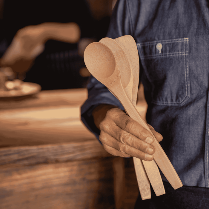 A person in a denim shirt holds three Bambu Kitchen Basics bamboo cooking utensils in a warmly lit kitchen.