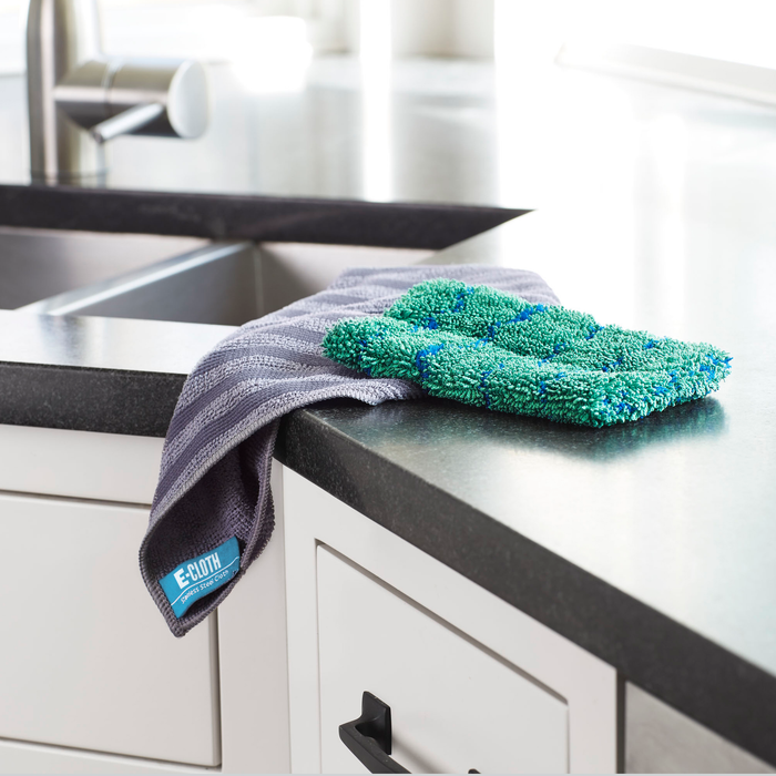 A stainless steel sink area displays an E-Cloth branded Kitchen Dynamo Microfiber Scrubber and an eco-friendly sponge. Bright natural light from a nearby window highlights the green textured cleaning pad.