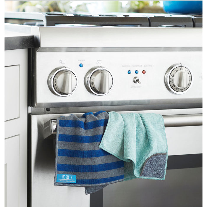 A stainless steel stove with dual knobs and a digital clock catches the eye, accompanied by two microfiber cleaning cloths—one striped blue, one light blue—from the E-Cloth Kitchen Pack hanging neatly from its handle. A white kitchen cabinet partially appears on the left.