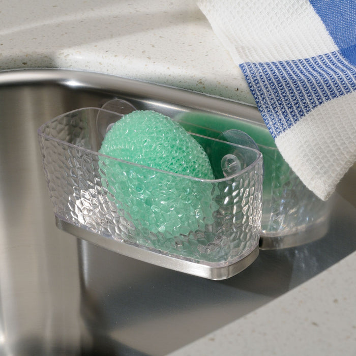The iDesign Kitchen Sink Sponge Suction, crafted from recycled plastic, holds a green scrub sponge in its clear textured holder inside the sink. A white and blue checkered towel peeks out alongside it on a light countertop.