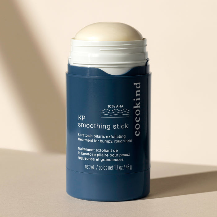 The dark blue Cocokind KP Smoothing Stick with a white cap stands upright against a beige background, highlighting 10% AHA for effective keratosis pilaris treatment and smoothing rough skin, with shadows casting softly to the right.