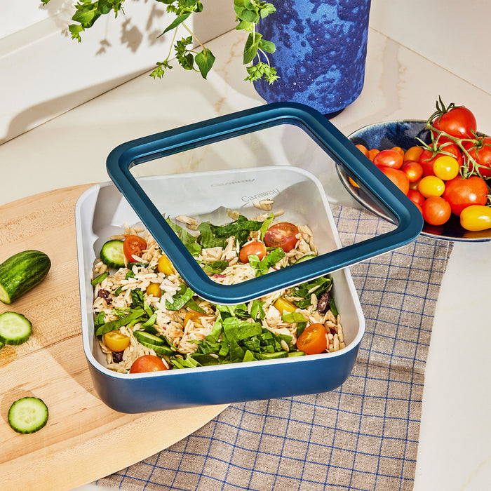 A Large Glass Food Container by Caraway, featuring a transparent lid and blue base, holds a salad of spinach, cherry tomatoes, and orzo pasta. Positioned next to sliced cucumbers on a cutting board and a blue bowl of tomatoes, this non-toxic container keeps your meal fresh and safe.