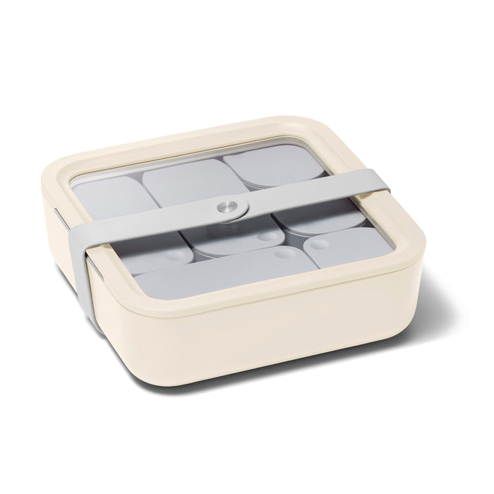 The Caraway Large Glass Food Container is a square, cream-colored storage solution with a transparent lid and gray strap. It includes gray dividers for compartmentalization, ensuring non-toxic and perfectly organized contents.