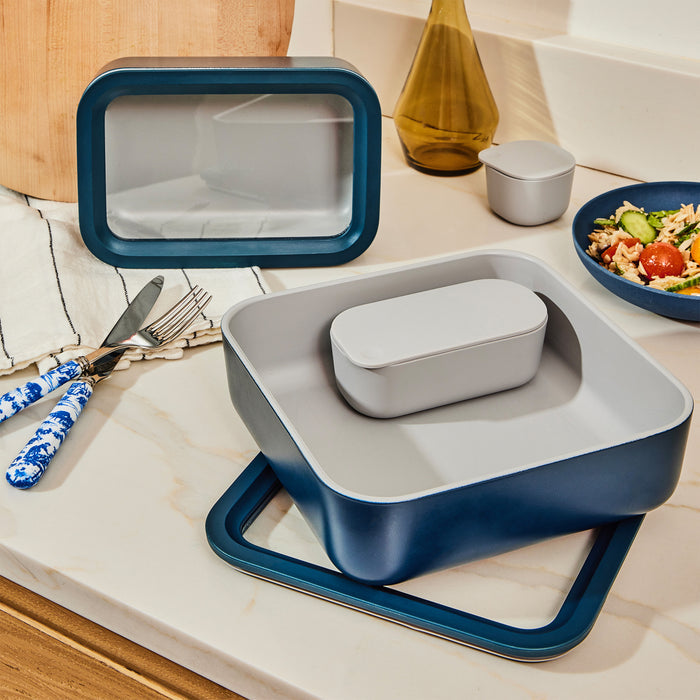 Kitchen countertop featuring Caraways large glass food container, non-toxic and available in various shapes. Includes a small salad bowl, utensils with blue patterned handles, and a gold decorative vase. White and wood accents create a cozy atmosphere.