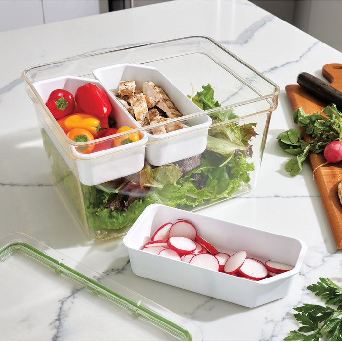 The iDesign Large Produce Bin with 3 Dividers, made from recycled plastic, holds fresh salad greens with compartments for bell peppers, sliced chicken, and radishes. Its lid rests on a marble countertop beside a cutting board holding radishes and herbs.