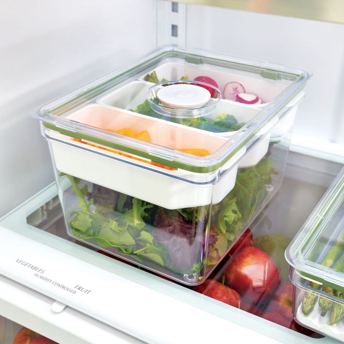 The iDesign Large Produce Bin with 3 Dividers, made from recycled plastic and featuring a white lid, stores leafy greens, sliced bell peppers, broccoli, and radishes in a refrigerator. Several apples sit below.