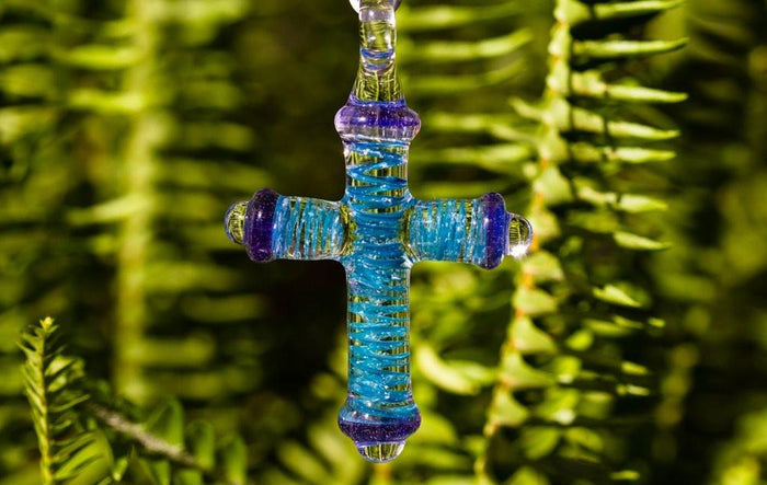 Peacock blue and transparent purple glass cross pendant infused with cremation ashes, shown outdoors among green ferns.