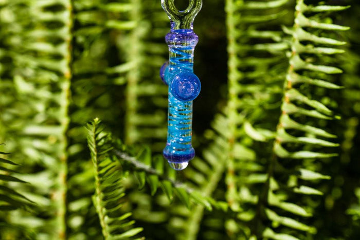 Blue and purple glass cross pendant infused with cremains, shown from the side with decorative cap against greenery.