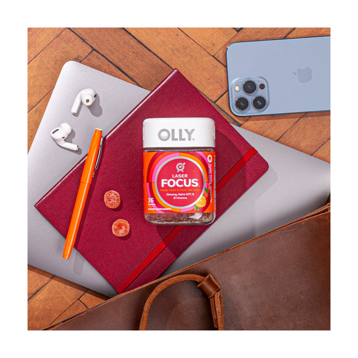 A top view of a workstation showcases a container of OLLY Laser Focus gummies, featuring Alpha GPC and Ginseng, on a red notebook. Nearby are a silver laptop, orange pen, two sliced gummies, white wireless earbuds, blue smartphone, and part of a brown leather bag.