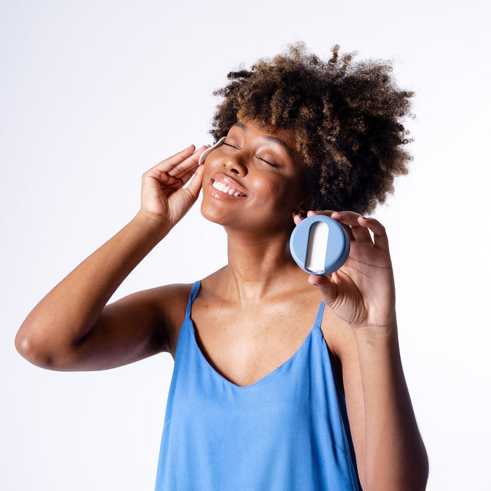 In a plain white setting, someone in a blue camisole smiles while using eco-friendly skincare on their cheek. They hold LastRound, a sustainable product by LastObject, in a round container with a lid.
