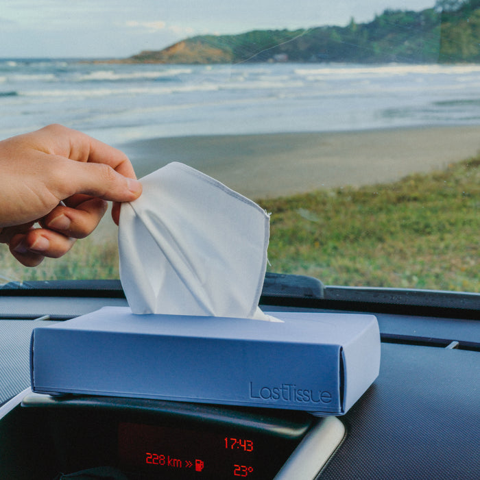 A hand pulls a white cloth from an eco-friendly LastTissue Box by LastObject on a car dashboard. Through the windshield, theres a scenic beach and ocean view with distant hills, while the dashboard displays 228 km and 17:43.