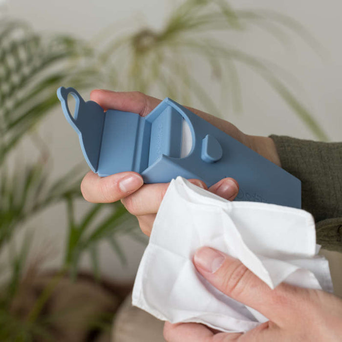 A person holds a blue plastic handheld device by LastObject, likely a cleaning tool, in one hand and a white organic cotton tissue called LastTissue in the other. A leafy plant in the background emphasizes an eco-friendly ambiance.