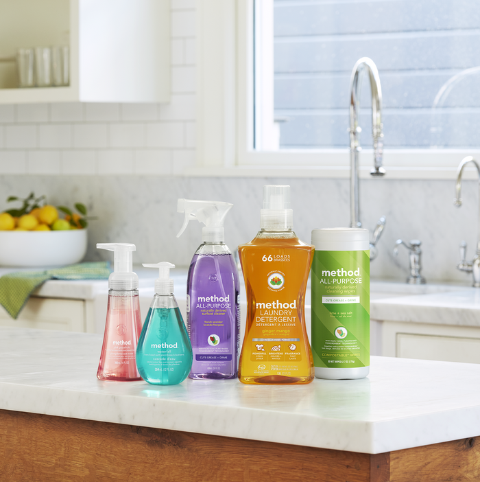 Five method cleaning products, like foaming hand wash and all-purpose cleaner, sit on a marble countertop next to the method Laundry Detergent Pour. A bowl of lemons enhances this kitchen scene that champions cruelty-free cleaning products.