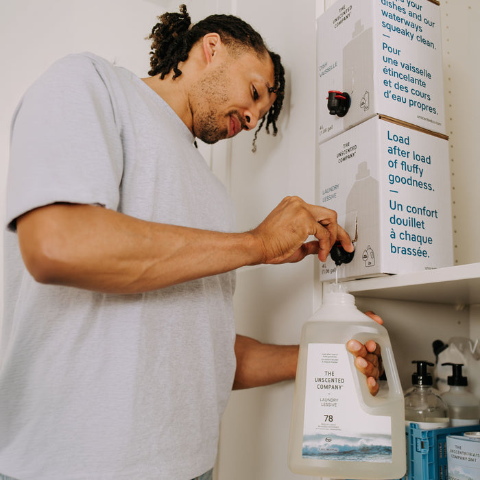 A person refills a large bottle of The Unscented Company Laundry Detergent, which is hypoallergenic and free of fragrance and dye, from a wall-mounted dispenser in the laundry room.