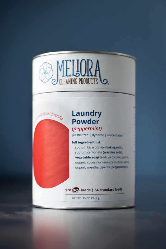 A cylindrical container of Meliora Laundry Powder in peppermint scent is shown on a blue background, showcasing its eco-friendly features: plastic-free, dye-free, synthetic fragrance-free, with a full ingredient list.