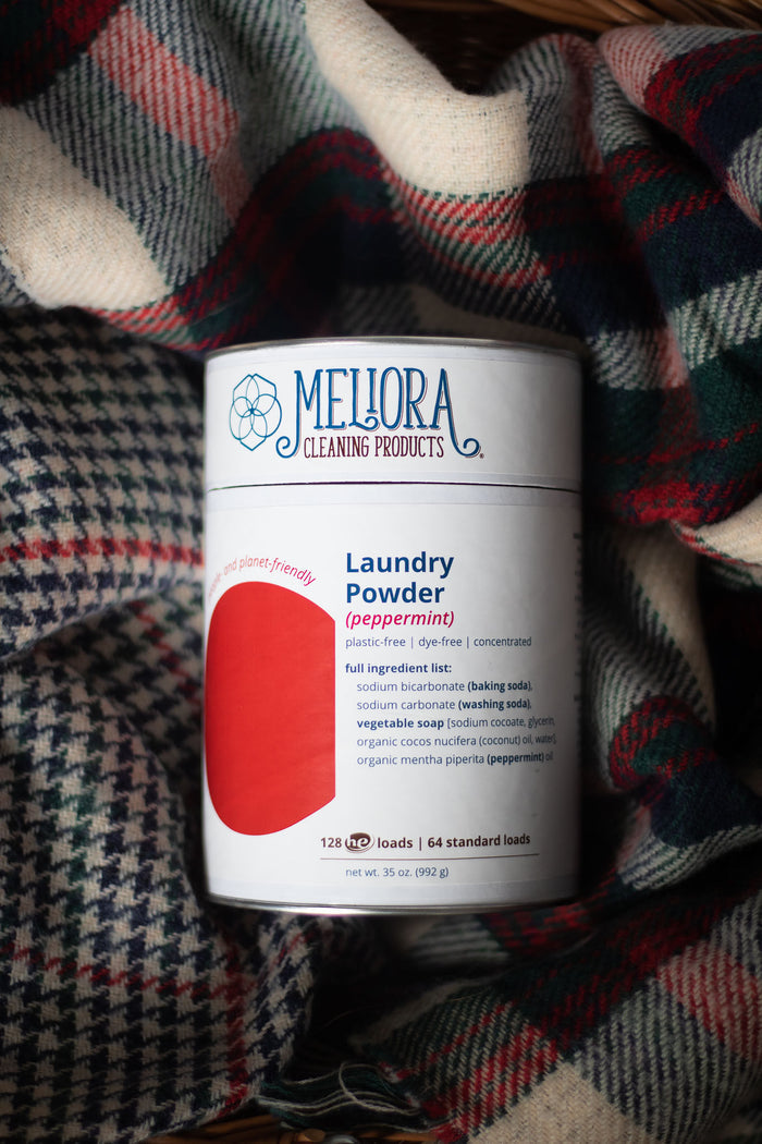 A canister of Meliora Laundry Powder (peppermint) sits on a plaid fabric, showcasing its eco-friendly features: plastic-free, synthetic fragrance-free, with concentrated ingredients like baking soda and peppermint oil.
