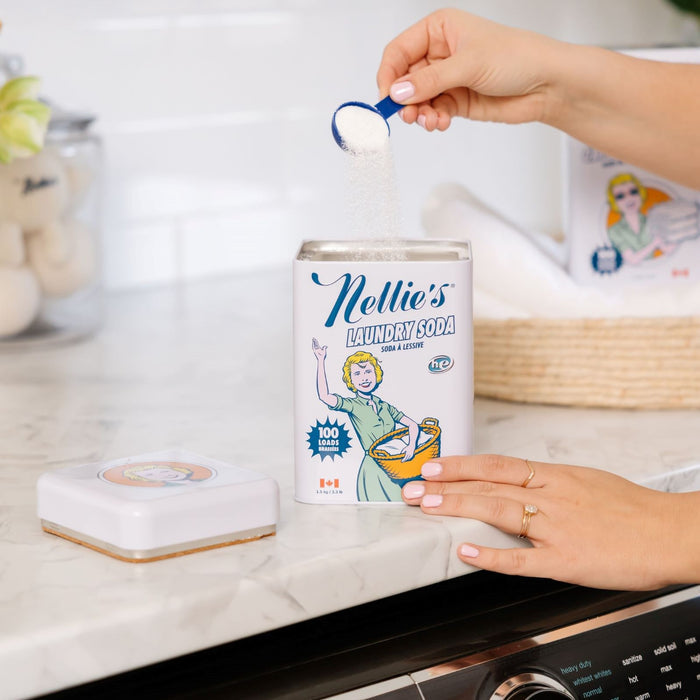 Someone is scooping Nellies Laundry Soda from a tin on a kitchen counter. The tin features vintage-style graphics with a woman in a green dress, and the matching lid is nearby, enhancing the nostalgic charm.