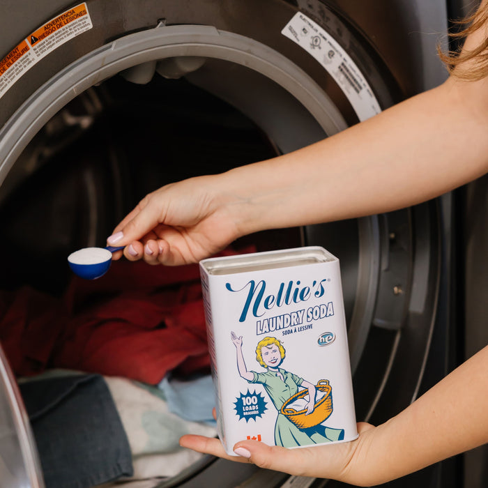 Someone uses Nellies Laundry Soda, scooping some powder into a front-loading washer. The container features a vintage-style illustration of a woman with a laundry basket, adding to the appeal of this effective detergent.