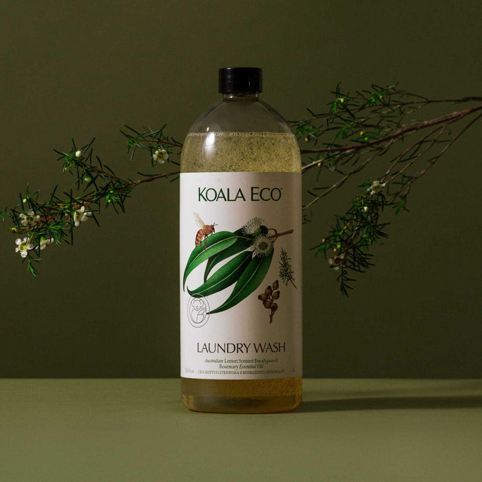A bottle of Koala Eco Laundry Wash - Lemon-Scented Eucalyptus & Rosemary with a botanical label sits on a green surface, paired with a leafy branch and small white flowers, evoking fresh Australian scents.
