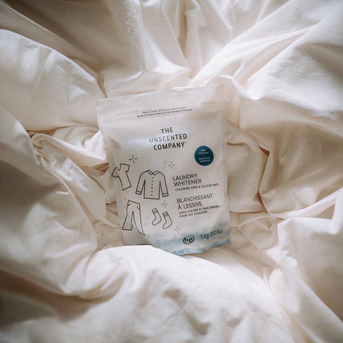 A package of The Unscented Company Laundry Whitener + Brightener, a colour-safe natural bleach alternative, rests on soft white fabric. The packaging displays simple clothing illustrations and bilingual English-French text.
