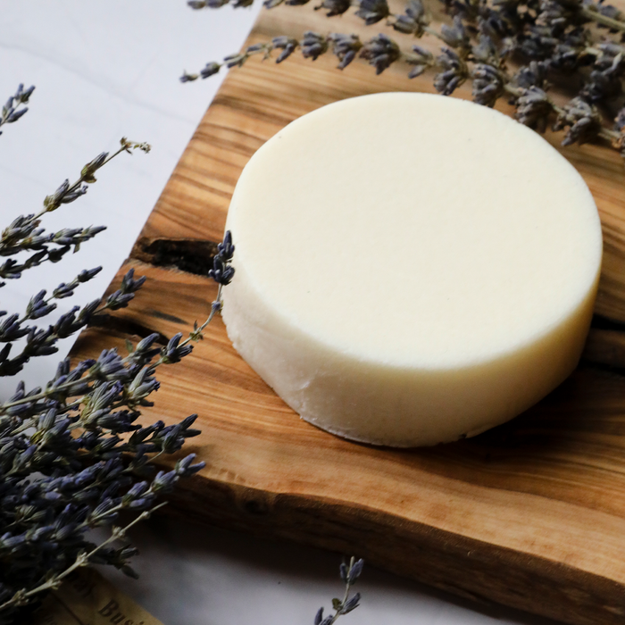 A round Lavender Body Butter Bar from Spinster Sisters Co. sits on a rustic wooden board with dried lavender sprigs. The off-white bar contrasts the muted purples, creating a calming, natural aesthetic that highlights its soothing ingredients of lavender essential oil.