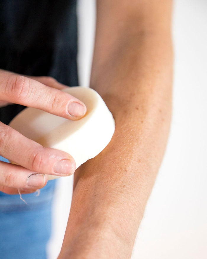 A person rubs a Spinster Sisters Co. Lavender Body Butter Bar, infused with Fair Trade shea butter, on their forearm. Their freckled skin glows softly against the blurred background, contrasting with their dark shirt and blue pants in this moment of serene vegan skincare indulgence.
