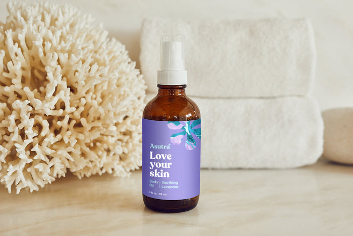 A brown bottle labeled Asutra Lavender Body Oil rests on a marble surface, complemented by coral and folded white towels, emphasizing the soothing benefits of lavender in this natural skincare product.